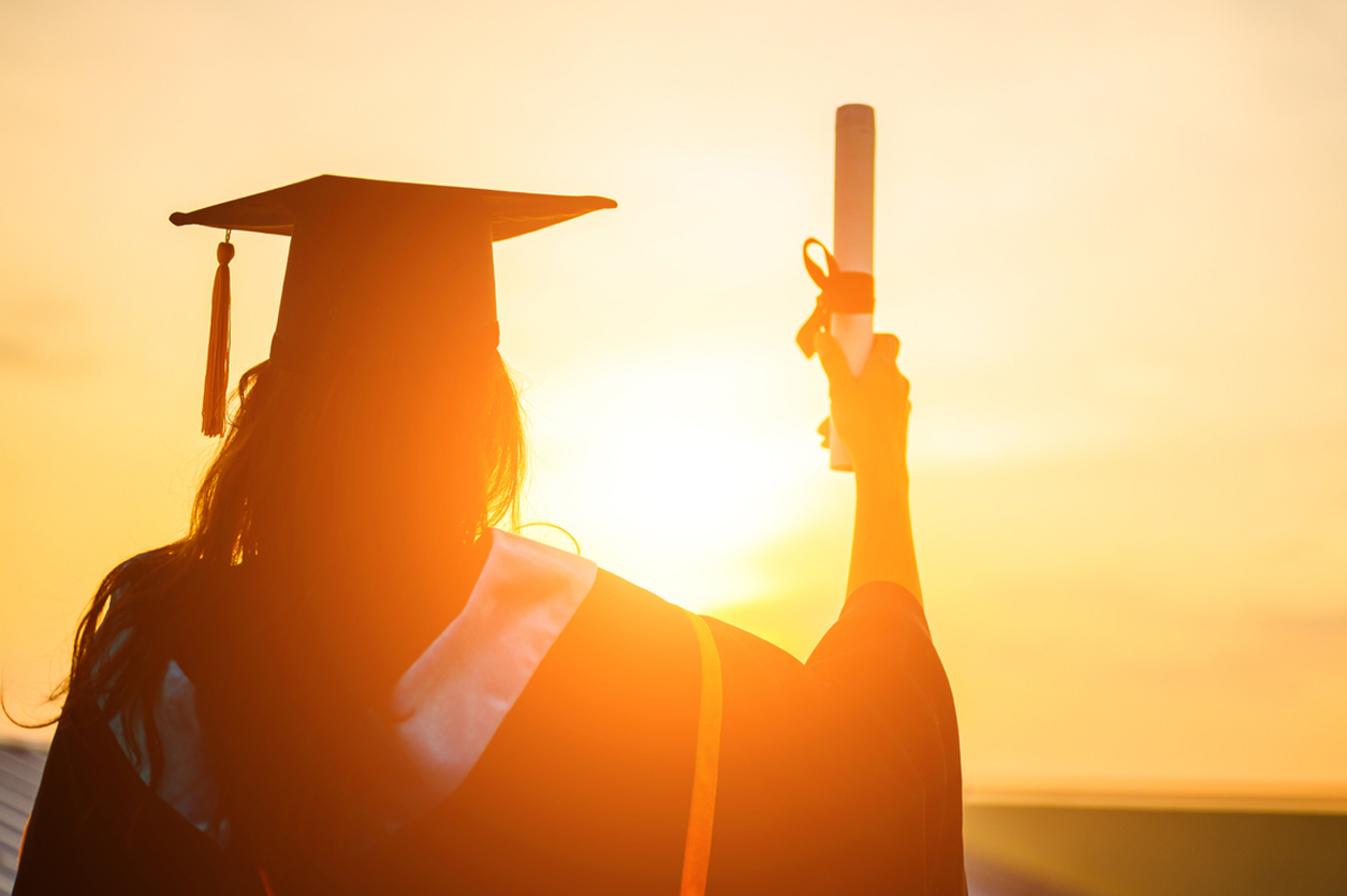 Silhouette of a woman in a graduate hate holding diploma up backlit by a sunset